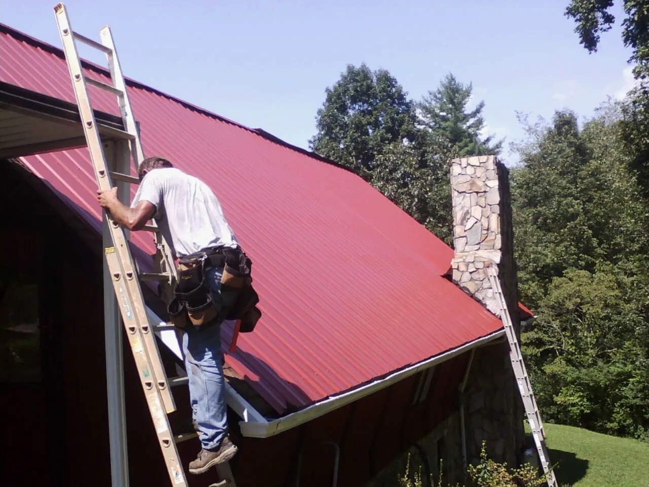 Roofer on a ladder beside a red metal roof and stone chimney — Soffit & Fascia Repair by Roofing Brothers in Forks Of Salmon